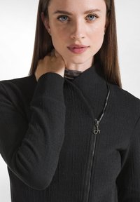 Giacca nera con zip, realizzata in tessuto testurizzato, colletto alto, maniche lunghe e dettaglio della cerniera argentata. Primo piano del design frontale.