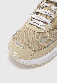 Beige athletic shoe featuring a mesh upper, rubber sole, white laces, and subtle grey accents. Rounded shape with textured details.