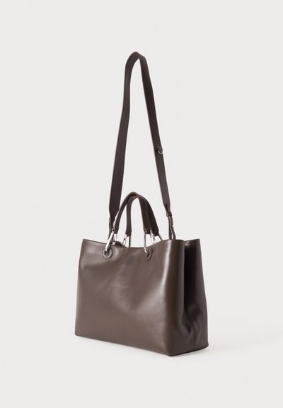Sac cabas en cuir marron foncé avec deux poignées courtes et une longue bandoulière réglable, debout sur un fond clair.