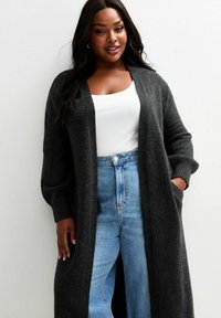 Un long cardigan gris foncé en tissu doux, avec un devant ouvert, des épaules tombantes et des poches latérales, associé à un haut blanc et un jean bleu clair.