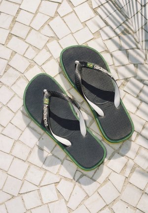 Para ciemnoszarych i zielonych klapek Havaianas na białej, wzorzystej podłodze kafelkowej z cieniem liści palmy.