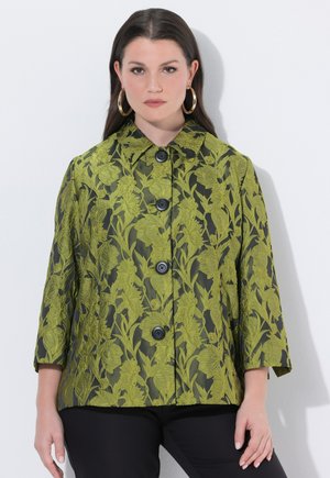 Žena oblečená v zelené bundě s květinovým vzorem, velkými černými knoflíky a zlatými kruhovými náušnicemi, stojící před bílým pozadím.