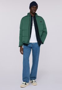 Chaqueta acolchada verde, camisa de mezclilla, camiseta blanca y jeans azules. Zapatillas blancas con acentos verdes. Gorro de punto. Pose de pie.