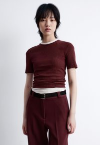 Burgundy ribbed t-shirt with short sleeves and contrasting white neckline, paired with dark trousers and a black belt, featuring a smooth texture.