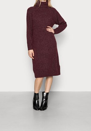 Robe col roulé en tricot bordeaux avec manches longues, détails côtelés à l'ourlet, associée à des bottines noires. Texture lisse, longueur mi-mollet.