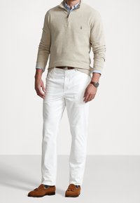 Pull en maille beige avec un col zippé mi-haut, associé à un pantalon blanc et des mocassins en daim marron, présentant un design classique et une texture lisse.