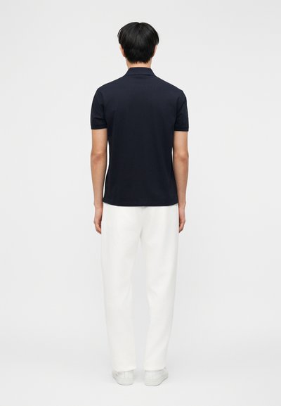 Άνδρας που στέκεται πλάτη φορώντας μπλε κοντομάνικο πόλο, λευκό παντελόνι και λευκά αθλητικά παπούτσια μπροστά σε απλό φωτεινό φόντο.