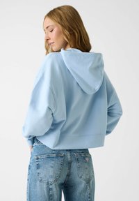 Sweat à capuche court bleu clair avec poche frontale et cordon de serrage à la capuche, associé à un jean denim bleu délavé avec un petit logo sur la poche arrière.