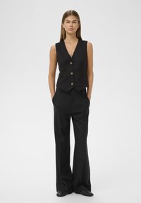 Black tailored vest with a V-neck, five buttons, and side pockets, paired with wide-leg black trousers in a smooth fabric.