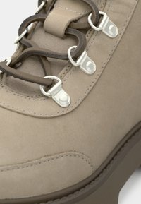 Beige semsket ankelstøvler med metallhardware, brune snørebånd og en teksturert såle. Buet søm gir en detaljert aksent til designet.