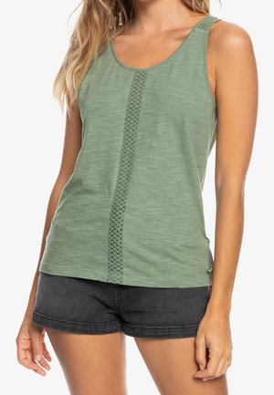 Frau trägt ärmelloses grünes Tanktop mit vertikalem Spitzenstreifen und schwarze Jeansshorts, steht mit entspannt an den Seiten hängenden Armen.