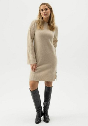 Kvinna med långt blont hår iklädd en beige stickad klänning med mönstrade ärmar och svarta läderstövlar som går upp till knäna, stående mot en enfärgad bakgrund.