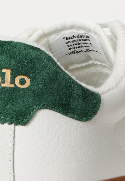 Vit sneaker med en grön mocka detaljer vid hälen, guldpräglad logotyp och en blank yta. Snören och texturen är detaljerade upptill.