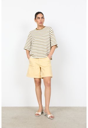 Vrouw staat met handen in de zakken, draagt een gestreept oversized t-shirt, beige korte broek en witte sandalen met hak, tegen een effen witte muur.