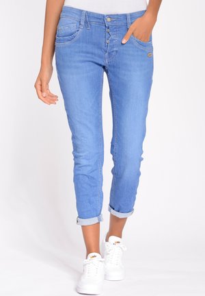 Vrouw draagt lichtblauwe opgerolde jeans met knoopsluiting en witte Nike sneakers, staat met één hand in de zak.