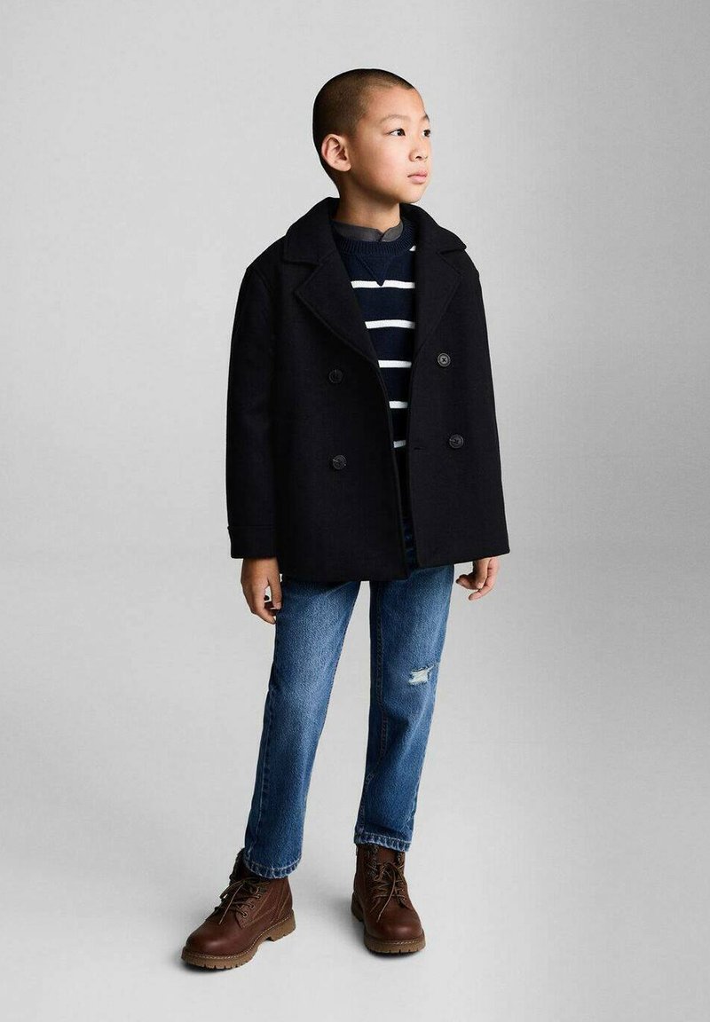 Svart dubbelknäppt sjömanjacka över en marinblå randig tröja, i kombination med blå rivna jeans och bruna snörboots.