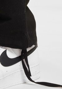 White leather sneaker with black accents, featuring a textured finish, thick sole, and black laces. Pants are black and slightly frayed at the hem.