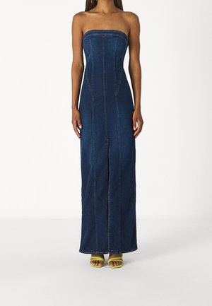 Woman wearing strapless dark blue denim maxi dress with front seam details and slit, paired with yellow open-toe heels, standing against white background.