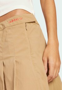 Beige skirt by Adidas with a pleated design, featuring a zip closure and embroidered logo in red on the waistband. Smooth texture.