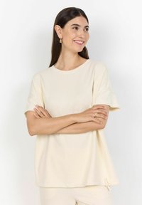 Cremefarvet T-shirt med korte ærmer, rund halsudskæring, afslappet pasform og toggle-detalje ved kanten. Blødt, glat stof med minimal tekstur.