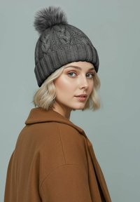 Gray knitted beanie with a patterned texture and a fluffy pom pom on top, worn by a person in a brown coat, against a light background.