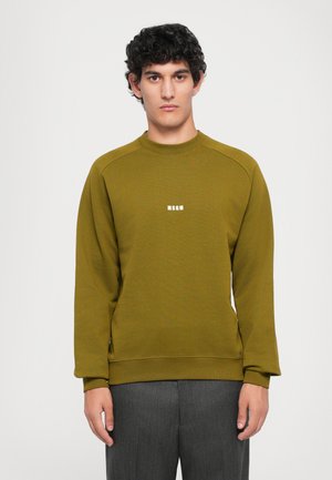 Bărbat cu păr creț și închis la culoare, îmbrăcat cu un hanorac MSGM verde măsliniu și pantaloni gri închis, stând în fața unui fundal deschis uniform.