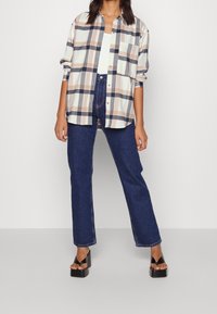 Geverifieerd overhemd in beige, marineblauw en roomkleur met knopen en een relaxte pasvorm. Gepaard met hoge taille donkere spijkerbroek en zwarte platformsandalen.