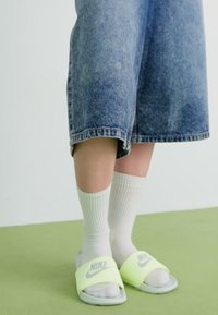 Nike-sandaler i fluorescerende gul med grå detaljer, brukt med hvite knelengdesokker, parret med blå denimshorts. Grønn gulvbakgrunn.
