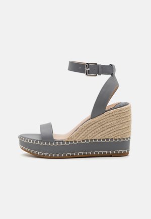 Sandale à talon en coin grise avec bracelet de cheville, bout ouvert et plateforme en jute tissée cousue sur le bord de la semelle, sur fond blanc.