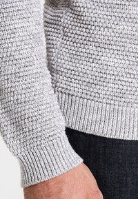 Grå stickad tröja med en ärm som har ett texturerat mönster. Ribbad mudd med en kontrasterande slät kant, i kombination med mörka jeans.