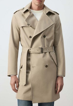 Hombre con gabardina beige de doble botonadura con cinturón en la cintura y hombreras sobre un suéter blanco y vaqueros azules.