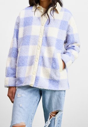 Person iført en blå og hvid ternet fleecejakke med knapper og lyse revnede jeans, hånd i lommen, mod ensfarvet baggrund.