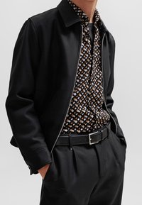 Zwarte jas met ritssluiting, gedragen over een patroonhemd met geometrische ontwerpen in bruin, beige en wit. Een zwarte riem maakt de look compleet.