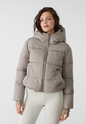 Veste coupe-vent beige avec capuche, présentant des coutures horizontales, une fermeture éclair et des poignets élastiques. Tissu lisse et léger.