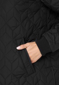 Hand med ljusrosa nagellack i fickan på en svart kviltad jacka med ribbad manschett.