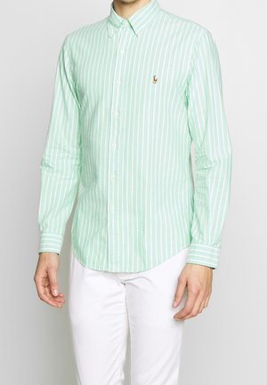 Hombre con camisa de botones de rayas verticales en verde claro y blanco, y pantalones blancos, contra un fondo claro y liso.