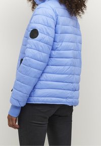 Persona con cabello rizado que lleva una chaqueta acolchada de color azul claro con un parche negro circular en la manga izquierda y pantalones negros.