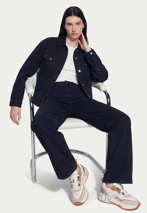 Jeune femme assise sur une chaise blanche, portant une veste en jean noire, un jean noir taille haute, un t-shirt blanc et des baskets blanches avec des semelles orange.