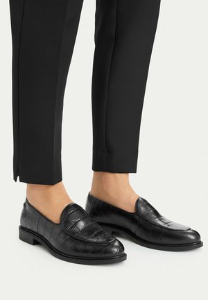 Black leather loafers with crocodile texture worn with black cropped trousers on bare feet.