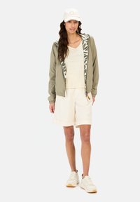 Lettere olivengrøn bomberjakke med en camouflageforet hætte, parret med en cremet top og afslappede cremede shorts, fuldendt med hvide sneakers.