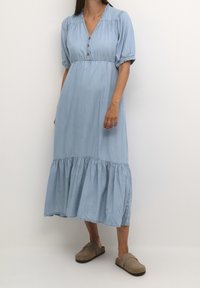 Kvinna iklädd en ljusblå knappradad midiklänning med puffärmar och beigea pjäxor, ståendes mot en enfärgad vit bakgrund.