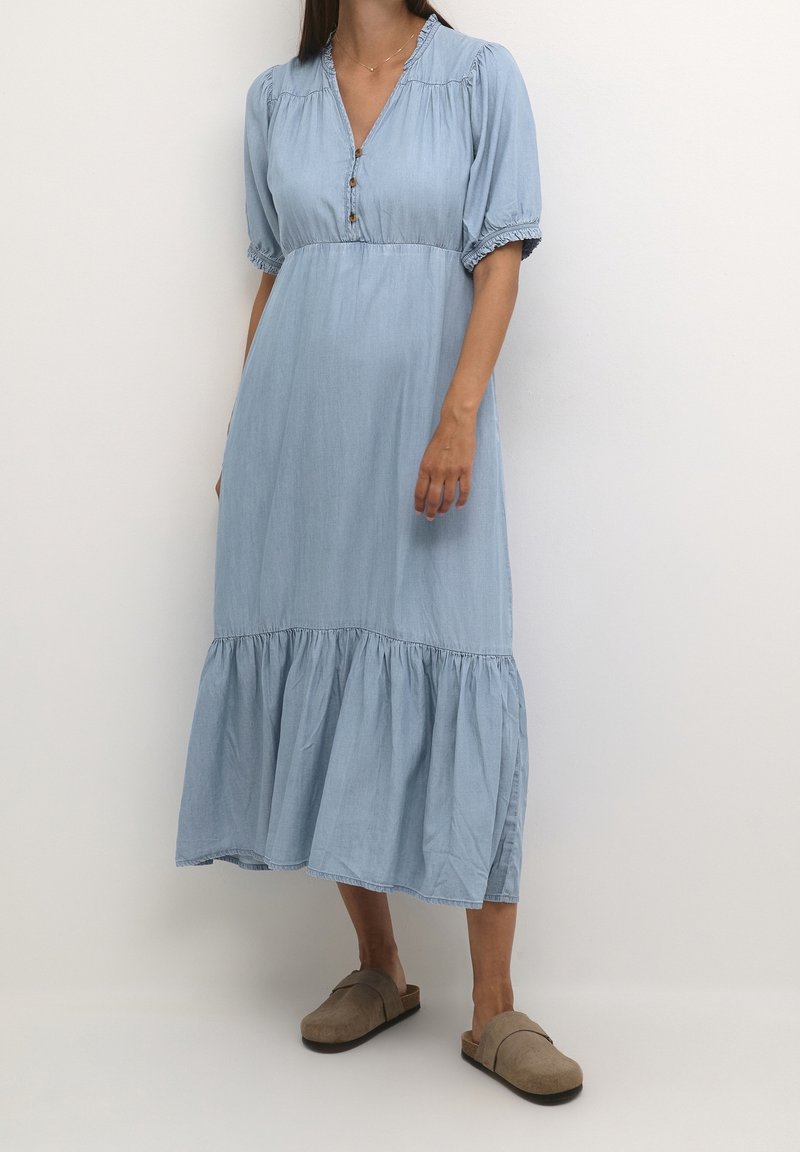 Kvinna iklädd en ljusblå knappradad midiklänning med puffärmar och beigea pjäxor, ståendes mot en enfärgad vit bakgrund.