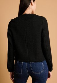 Čierna textúrovaná bunda s dlhými rukávmi, skráteným strihom a čistými líniami. Nosí sa s tmavomodrými džínsami, ktoré majú zadné vrecká.