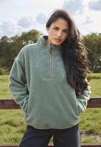 Sudadera de forro polar en verde apagado con un cuello de medio cremallera, que cuenta con un bolsillo de parche y una textura que recuerda a una tela suave y esponjosa.
