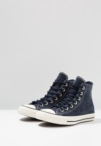 Námořnické semišové vysoké tenisky s bílým gumovým špičkovým krytem a podrážkou. Mají černé šňůrky a švy. Klasický design s kovovými průvleky.