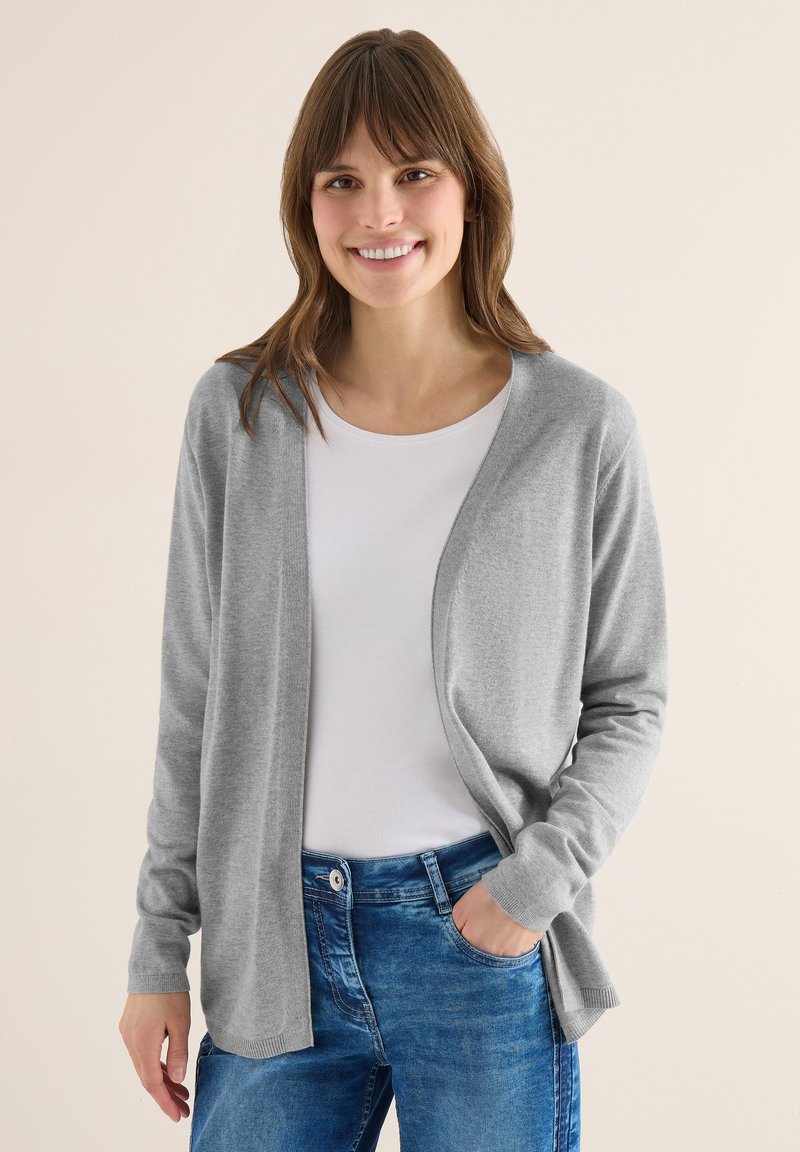Femme souriante portant un cardigan gris clair, une chemise blanche et un jean bleu, debout avec une main dans la poche devant un fond beige.