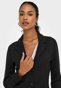 Femme aux cheveux tirés en arrière portant des boucles d'oreilles argentées en forme de créoles et un blazer noir, tenant le revers du blazer près de sa poitrine.