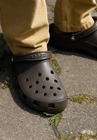 Bruna Crocs med ventilationshål, rund form, texturerad sula och justerbar hälrem, bärs med khakifärgade byxor på en asfalterad yta.
