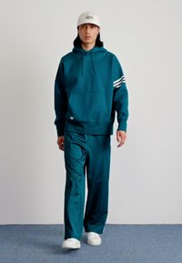 Teal hettegenser med frontlomme og snor, sammen med matchende bukser. Har tre hvite striper på skuldrene. Hvite joggesko og caps.