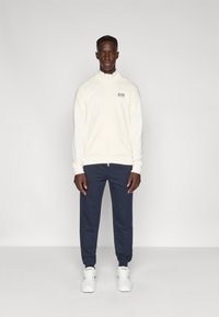 Φωτεινό μπεζ μπουφάν με φερμουάρ, ψηλό γιακά, ριμπ μανσέτες και λογότυπο, σε συνδυασμό με σκούρο μπλε παντελόνι. Το μοντέλο στέκεται μπροστά σε απλό φόντο.